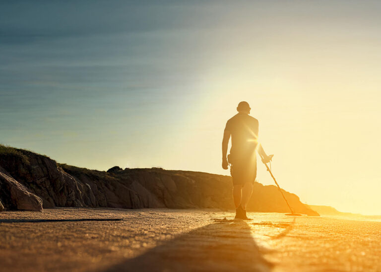 Nokta Legend 2 SMF Metal Detector Beach Detecting at Sunset Person using a Nokta Legend 2 SMF metal detector on a sandy beach at sunset with rocky cliffs.