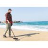 A bearded man in a plaid shirt and cargo pants uses a Nokta FINDX metal detector on a sandy beach with the ocean and rocky cliffs in the background.