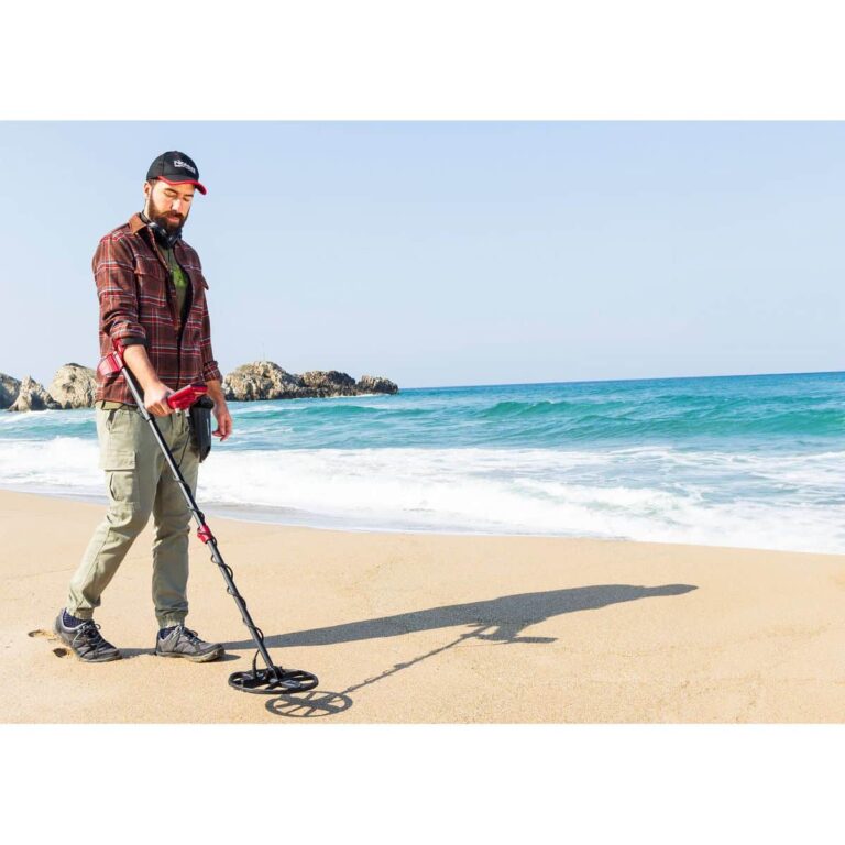 Beach Metal Detecting with Nokta FINDX A bearded man in a plaid shirt and cargo pants uses a Nokta FINDX metal detector on a sandy beach with the ocean and rocky cliffs in the background.