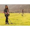 A woman and a man using Nokta FINDX metal detectors in a grassy field with mountains in the background.